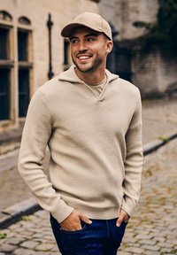 Beige gebreide pullover met een V-hals en ribbelstructuur, gecombineerd met een bijpassende beige pet, gedragen met donkere jeans. Op de achtergrond een straat met kasseien.