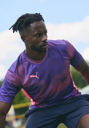 Paarse sportshirt met roze accenten en een stippenpatroon; korte mouwen en een ronde halsontwerp; model in actie op een sportveld.