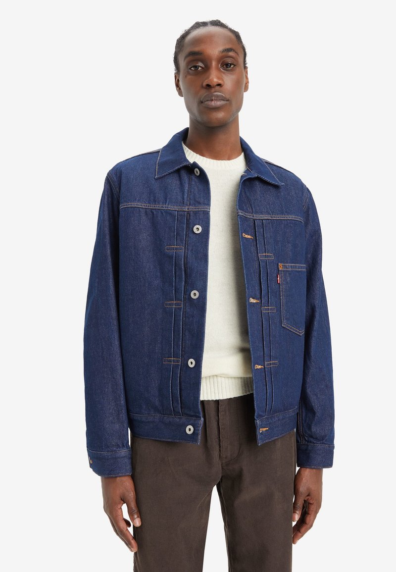 Dunkelblaue Jeansjacke mit Kragen, vorderer Knopfleiste, Brusttasche und kontrastierenden Nähten. Darüber getragen wird ein heller Pullover.