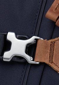 Navyfarbener Stoffbeutel mit Detail einer silbernen Metallschnalle und einem braunen Ledertrageriemen, der strukturiertes Nylon und kontrastierende Nähte zeigt.