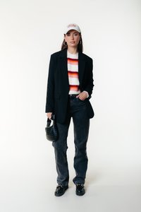 Woman wearing white baseball cap, striped sweater, black blazer, black jeans, black loafers, holding small black woven handbag against white background.