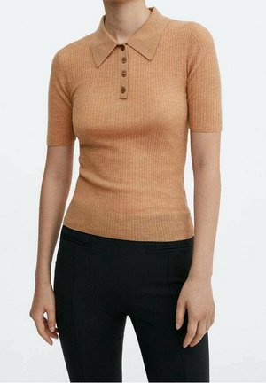 Kobieta ubrana w dopasowaną prążkowaną, krótką beżową koszulkę polo z czterema guzikami i czarne spodnie, stojąca na jednolitym tle.