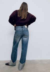 Breda ben, ljusblå denimbyxor med en blekt finish, med bakfickor och råa fållar. Kombineras med en vinröd tröja och grå skor.