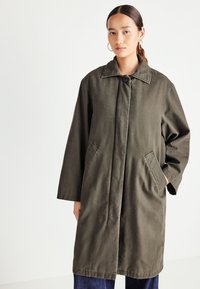 Woman wearing an olive-green long coat with front button closure and pockets, paired with dark blue jeans against a plain white background.