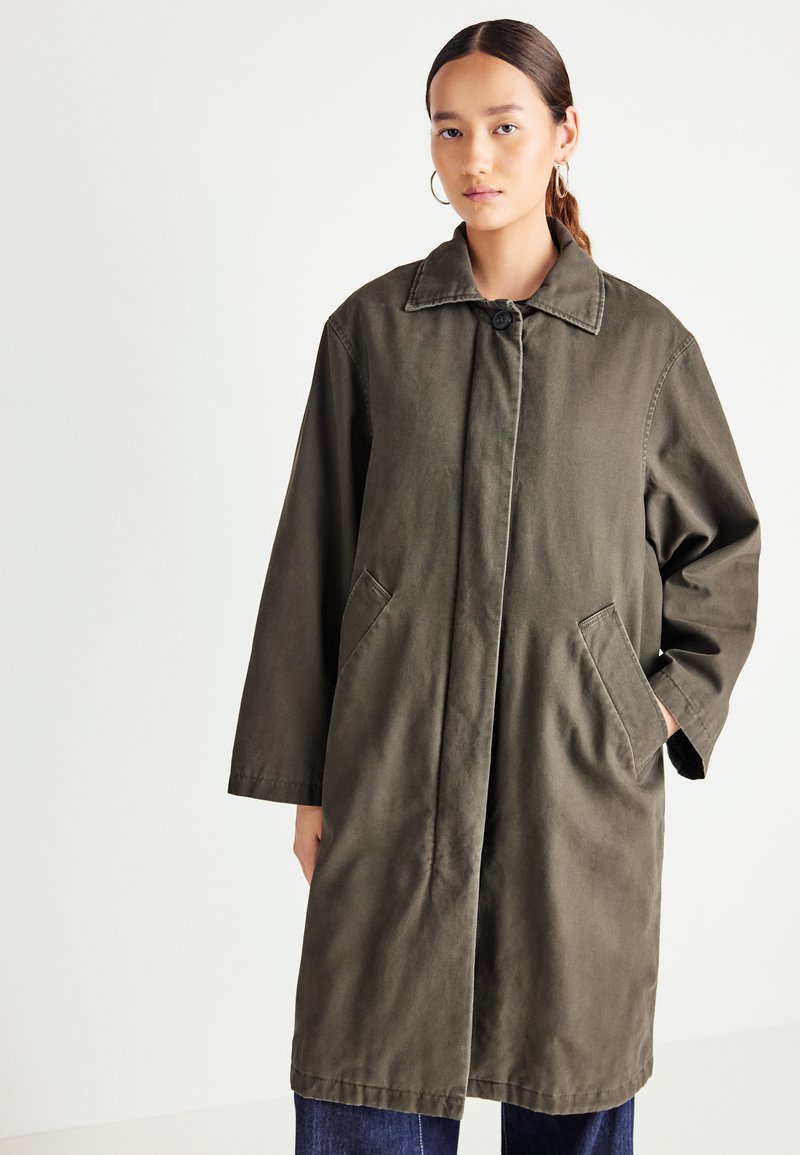 Woman wearing an olive-green long coat with front button closure and pockets, paired with dark blue jeans against a plain white background.