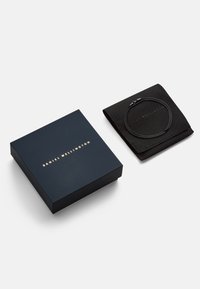 Sorte læderarmbånd i en foldet sort pose ved siden af en marineblå firkantet æske med guldskrift, begge med minimalistisch design.