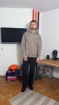 Light brown hoodie with front pocket and minimal branding, paired with dark pants and brown shoes, standing in a room with various decor.