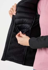 Mains ouvrant la poche intérieure d'une veste matelassée noire portée sur une chemise rose sur le torse d'une personne.