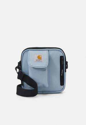 Borsa a tracolla Carhartt azzurro chiaro con tasca anteriore con patta, tasca con cerniera nera e tracolla nera regolabile su sfondo bianco.