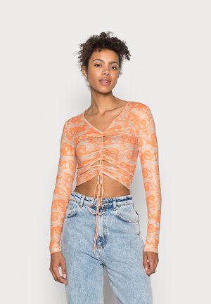 Top cropped de manga longa em tecido laranja transparente, com um padrão floral, detalhe frente franzido e decote em V profundo. Combinado com jeans azuis.