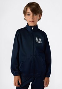 Granatowa kurtka z zamkiem błyskawicznym, wykonana z gładkiego materiału, z logo na lewej piersi, elastycznymi mankietami i bocznymi kieszeniami.