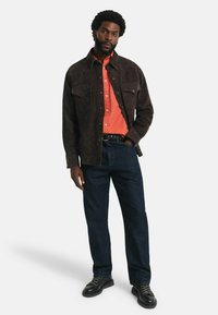 Braune Wildlederjacke über einem orangefarbenen Hemd, kombiniert mit dunkelblauen Jeans und schwarzen Schuhen. Merkmale sind zwei Brusttaschen und ein Knopfdesign.