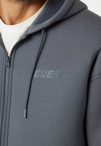Sudadera gris con cremallera y capucha con forro de felpa, que presenta un logotipo "GUESS" tonal, costuras dobles y cordones ajustables. Tejido suave.