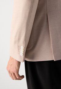 Detailný záber na ruku vedľa rukáva béžovo-kostkovaného blejzra so štyrmi bielymi gombíkmi, ktorý je skombinovaný s čiernymi nohavicami.