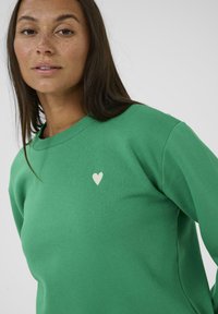 Grüner Sweatshirt mit rundem Halsausschnitt und langen Ärmeln, mit einem kleinen weißen Herz, das auf der Brust gestickt ist. Weicher, strukturierter Baumwollstoff.