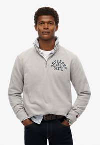 Sudadera de color gris claro con cremallera hasta el cuarto, con un logo en navy, llevada sobre una camisa de rayas blanca y azul. Unos jeans oscuros y un reloj de plata completan el conjunto.