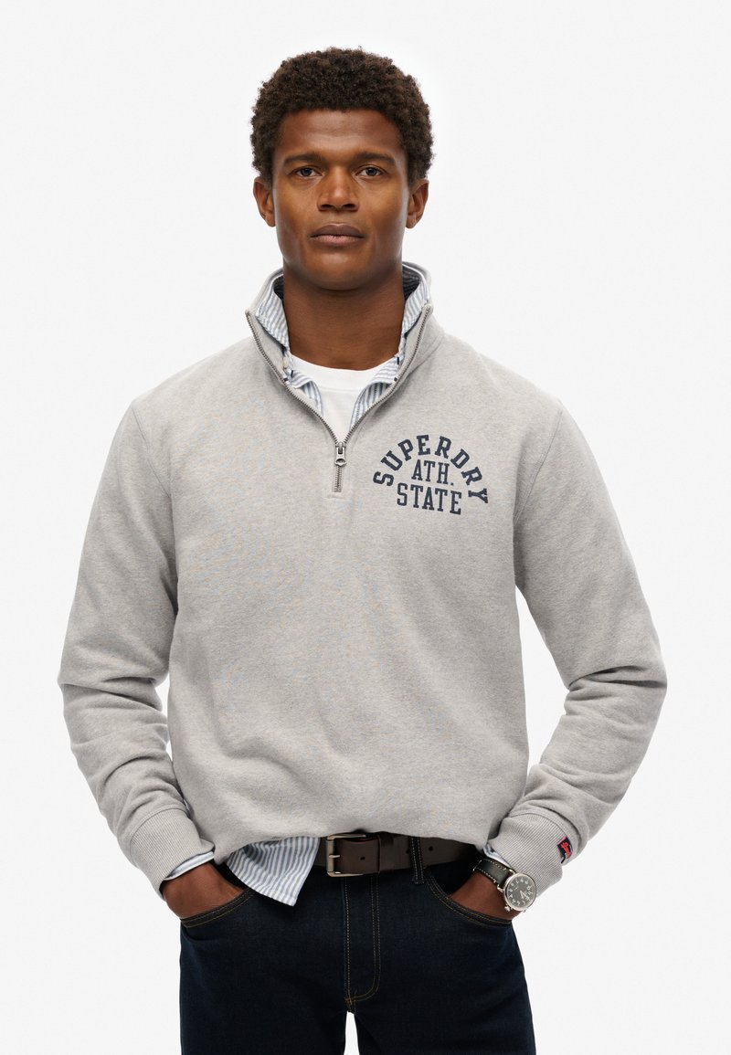 Sudadera de color gris claro con cremallera hasta el cuarto, con un logo en navy, llevada sobre una camisa de rayas blanca y azul. Unos jeans oscuros y un reloj de plata completan el conjunto.