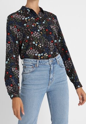 Femme portant un chemisier noir à manches longues à motifs floraux, rentré dans un jean taille haute bleu clair, les mains relâchées le long du corps, sur un fond uni.