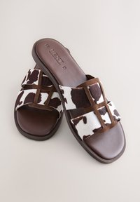 Braune Pantoletten mit Kuhfellmuster und weißen Akzenten. Weiche Textur, flache Sohle und überkreuztes Riemendesign für mehr Komfort.