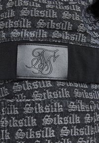 Tessuto nero con un motivo testurizzato caratterizzato da testo "Siksilk" ripetuto. Include un'etichetta in pelle nera liscia con iniziali in rilievo.