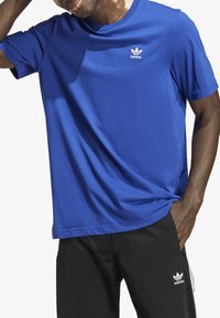 Camiseta de algodón azul con un cuello redondo clásico y mangas cortas. Presenta el logo blanco de Adidas en el lado izquierdo del pecho. Textura suave.