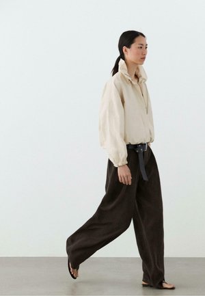 Frau geht, trägt eine beige gesteppte Jacke, lockere dunkelbraune Hose mit schwarzem Gürtel und schwarze Flip-Flop-Sandalen vor einem schlichten weißen Hintergrund.