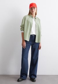Camicia a righe verde e bianca sopra una t-shirt bianca, abbinata a jeans a gamba larga blu scuro e un cappellino da baseball rosso. Scarpe nere.