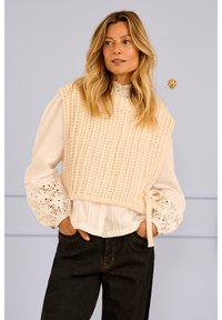 Suéter de punto color crema con nervaduras texturadas, superpuesto sobre una blusa blanca con puños de encaje floral. Vaqueros oscuros con detalles visibles de costura.