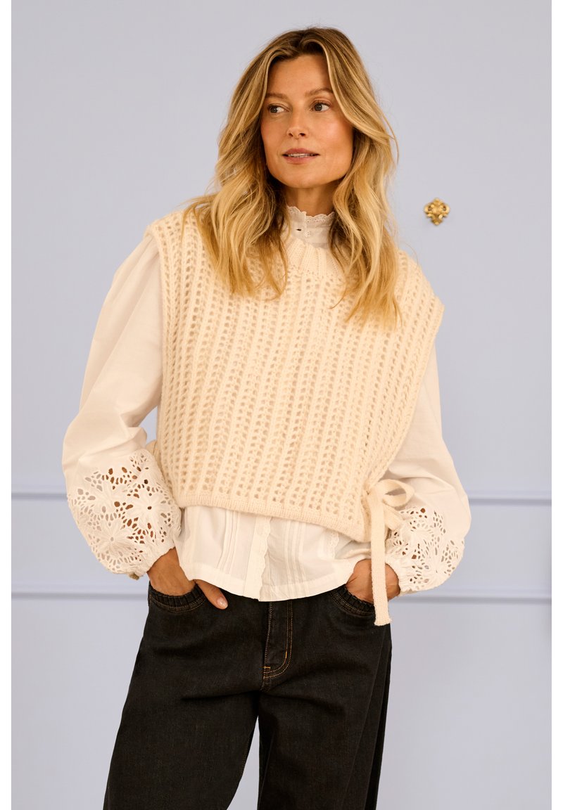 Suéter de punto color crema con nervaduras texturadas, superpuesto sobre una blusa blanca con puños de encaje floral. Vaqueros oscuros con detalles visibles de costura.