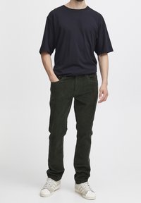 Zwart katoenen t-shirt met een losse pasvorm, gecombineerd met donkergroene ribfluwelen broek. Sneakers hebben lichte grijze en witte accenten.
