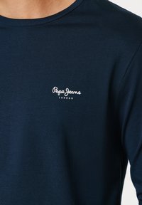 Marineblå langærmet skjorte med lille hvid "Pepe Jeans London"-logo på venstre bryst, båret af en person.