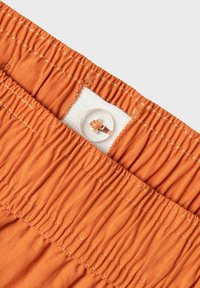 Ceinture en tissu orange avec une texture froncée, comportant une étiquette en tissu blanche et un détail de bouton transparent. Motif de couture simple le long du bord.