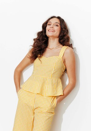 Jeune femme aux longs cheveux bouclés souriante, portant un top peplum sans manches à carreaux jaunes et blancs et un pantalon assorti, les mains dans les poches.