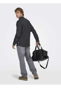 Sort lædertaske med to håndtag og aftagelig skulderrem; model iført mørk skjorte, grå jeans og brune sko.