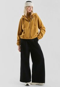 Braune Fleece-Zip-Hoodie mit Fronttaschen, kombiniert mit lockeren schwarzen Hosen und weißen Turnschuhen. Das Modell trägt ein weiches weißes Stirnband.