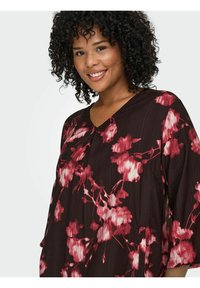 Smeđa cvjetna bluzica s dugim rukavima, s uzorcima crvenih i ružičastih cvjetova, V-izrezom i slobodnim krojem. Teksturirana tkanina dodaje dubinu.