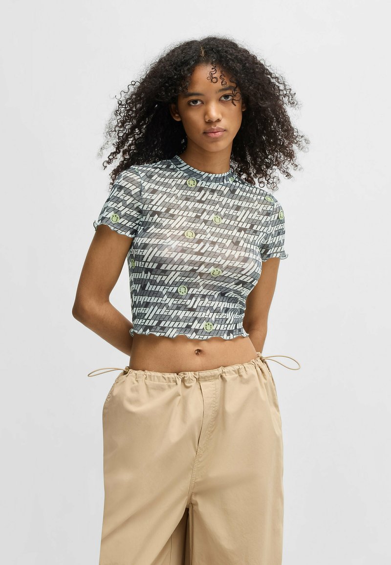 Short-sleeve, cropped top in sheer, textured fabric. Features a wavy hem and a pattern of alternating letters and smiley faces in gray and green.
