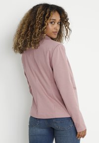 Blazer rose poudré à coupe cintrée, en tissu lisse et à manches longues. Doté d'un col à revers et de coutures arrière structurées. Associé à un jean bleu.