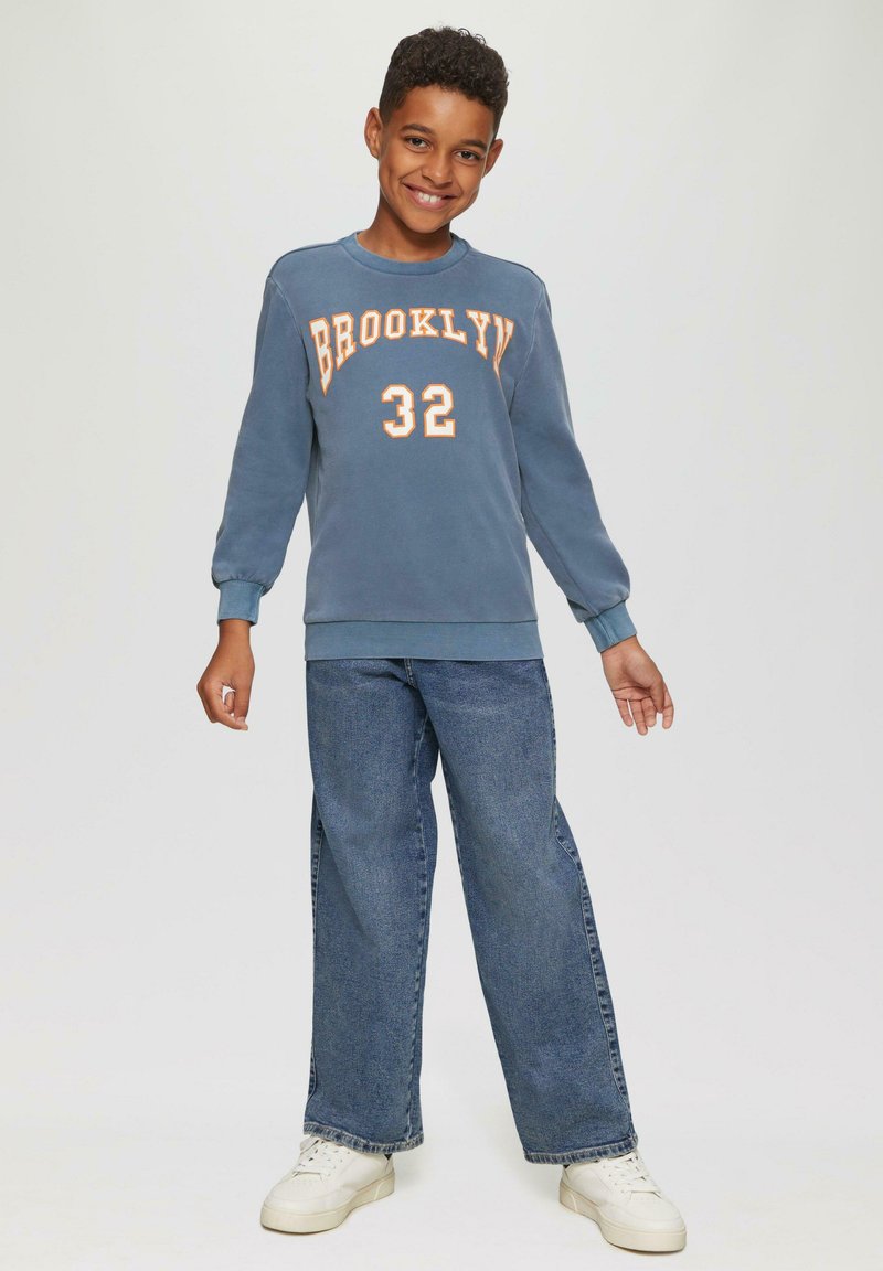 Blauwe sweatshirt met "BROOKLYN 32" in oranje, gecombineerd met wijde blauwe jeans en witte sneakers. Ontspannen pasvorm en lange mouwen.