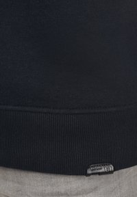Donkerblauwe sweatshirt met een geribde zoom, textuurstof en een klein zwart label met witte tekst in de linkerbenedenhoek.