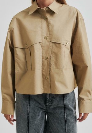 Femme portant une chemise beige boutonnée avec de grandes poches avant et un jean taille haute foncé avec des coutures visibles.