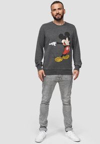 Grijze sweatshirt met lange mouwen met een groot bedrukt Mickey Mouse-ontwerp in rood, zwart en geel. Draagt het met lichtgrijze jeans en witte sneakers.