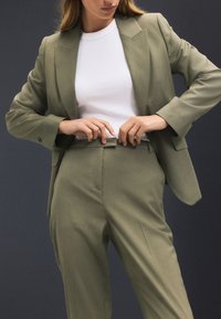Olivzaļš kostīms ar formētu žaketi un taisna griezuma biksēm, kombinēts ar baltu ribotu bezroku topu.