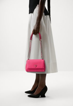 Bolso tejido rosa con un diseño estructurado, que incluye detalles de cadena y asa. Combinado con una falda blanca y zapatos de cuña negros.