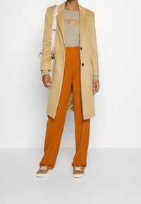 Cappotto in misto lana beige, top a coste oliva con apertura a chiave, pantaloni larghi color ruggine e sneakers marroni con motivo. Borsa beige con tracolla.