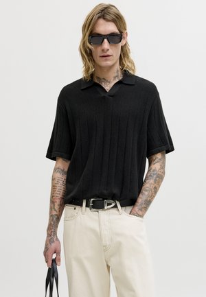 Νεαρός με μακριά μαλλιά και τατουάζ, που φοράει μαύρο ριμπ κοντομάνικο πουκάμισο, μπεζ παντελόνι, γυαλιά ηλίου και κρατάει χερούλια μαύρης τσάντας.