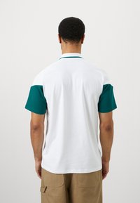 Kortermet polo skjorte med hvit kropp, turkise og beige accenter på ermene, en flat krage, og en teksturert stoffoverflate.