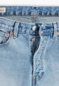 Světlé modré džíny z denimu se třemi viditelnými kovovými knoflíky na zapínání, oranžovým švem a měděnými nýty poblíž kapes.