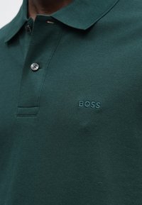 Zielona koszulka polo wykonana z teksturowanej tkaniny; posiada kołnierzyk, listwę na trzy guziki oraz wytłoczone logo "BOSS" na piersi.