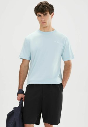 Joven con camiseta azul claro y pantalones cortos negros, sosteniendo una bolsa azul marino con una mano en un estudio blanco.